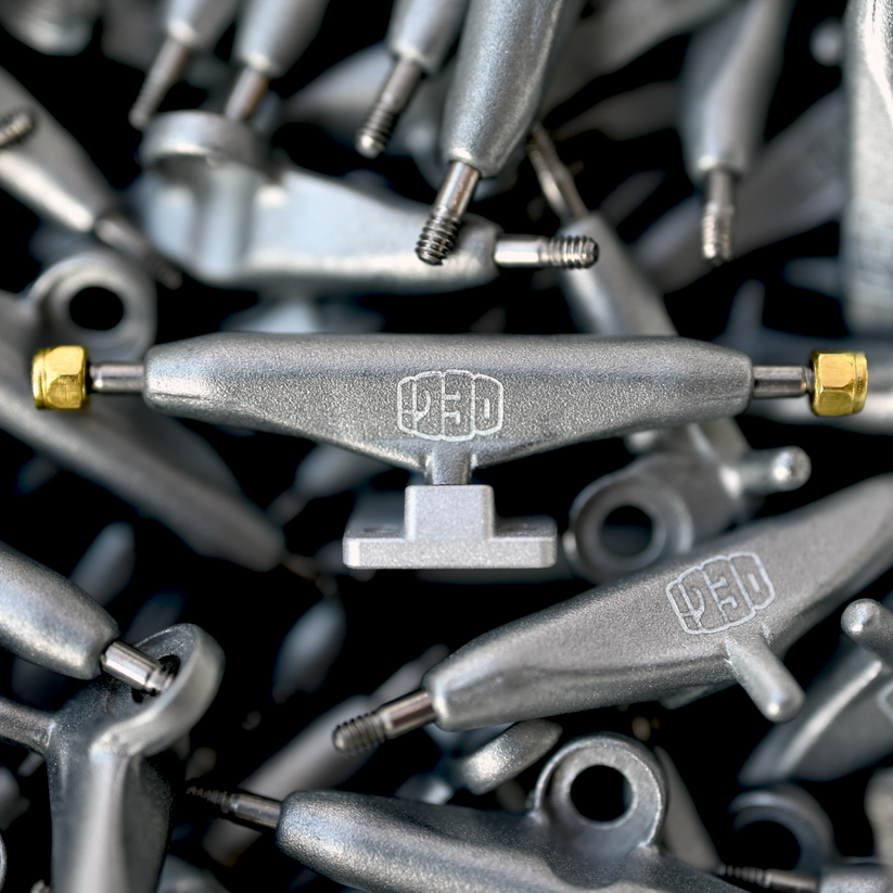 A close-up of the Deli Trucks Raw by tiny skateshop showcases a collection of glossy silver skateboard trucks. One is prominently centered with engraved text and gold hardware, while the background displays more trucks forming a metallic, textured pattern.